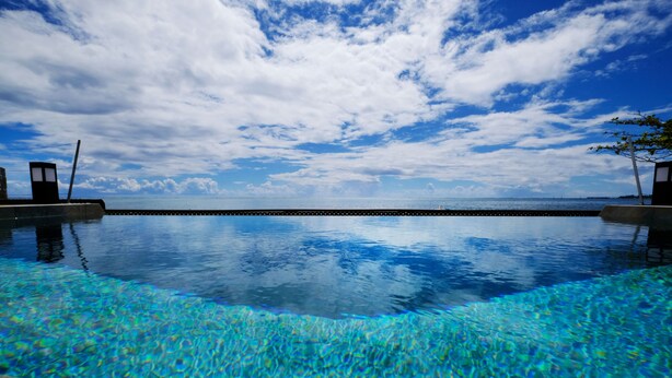 Céu azul límpido com nuvens, uma piscina calma com águas transparentes e um cais ao longe.