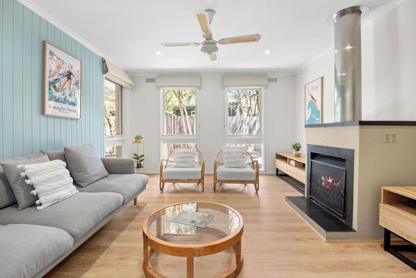 A living room with a grey sofa, two armchairs, a glass coffee table, a fireplace, and a ceiling fan.