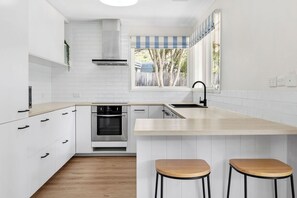 A modern kitchen with white cabinetry, a built-in oven, a stainless steel range hood, a large island with stools, and a window with a view of trees.