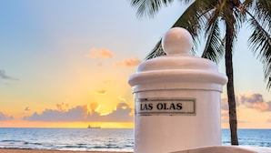 Escena de playa con un letrero blanco que dice 'LAS OLAS' y una palmera al fondo.