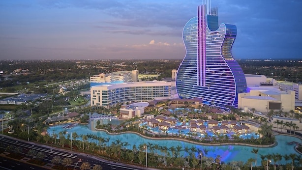 Un hotel grande con una edificación en forma de guitarra, varias piscinas y un área ajardinada alrededor.
