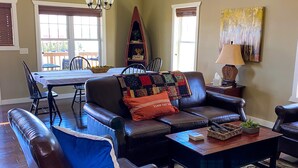 A living room with a sofa, coffee table, and a ceiling fan. There is a dining area with a table and chairs in the background. - Shoreline Cottage SD (Oacoma)