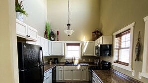 A kitchen with white cabinets, a black refrigerator, a microwave, and a coffee maker. There is a window with blinds and a hanging light fixture. - Shoreline Cottage SD (Oacoma)