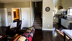 A spacious living area with a dark wood floor, a large window, and a view of the kitchen. - Shoreline Cottage SD (Oacoma)