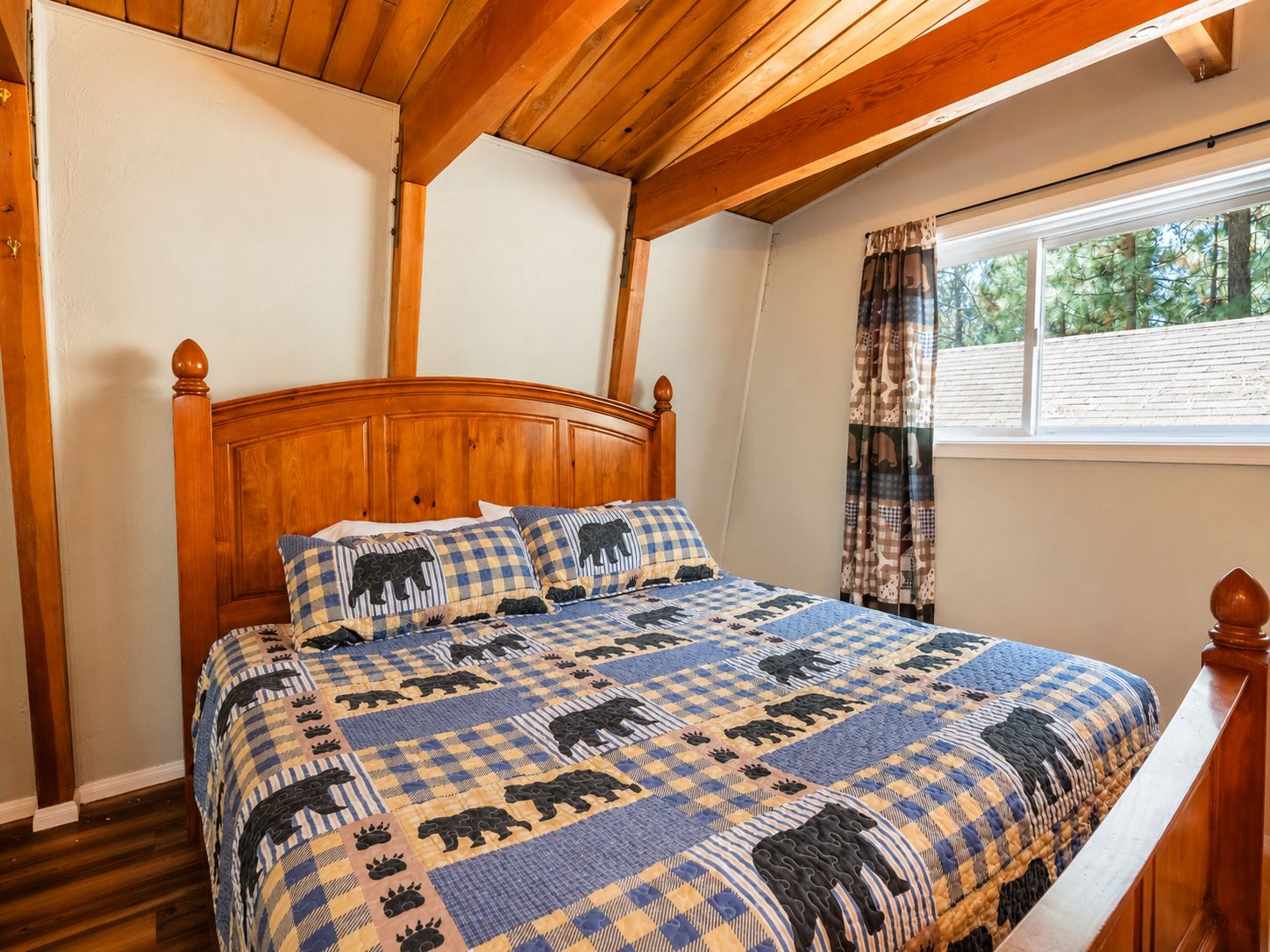 A bedroom with a wooden headboard, a checkered bedspread with animal prints, a window with curtains, and a wooden floor.