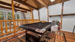A pool table in a room with wooden beams, a stone wall, and a view of trees outside. - Sherwood Pines - Pool Table, Fireplace, Fenced Yard (Big Bear City)