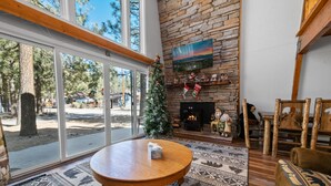 A cozy living room with a stone fireplace, a large window, and a Christmas tree. - Sherwood Pines - Pool Table, Fireplace, Fenced Yard (Big Bear City)