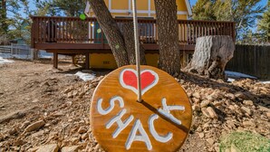 Ein Holzschild mit der Aufschrift "I ♥ SHACK", vor einem Hintergrund aus einem gelben Gebäude, Bäumen und einer Holzterrasse.