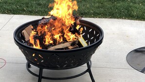 Ein schwarzer Feuerkorb für den Außenbereich mit Flammen, auf einer Betonfläche, mit einem Rasen im Hintergrund.