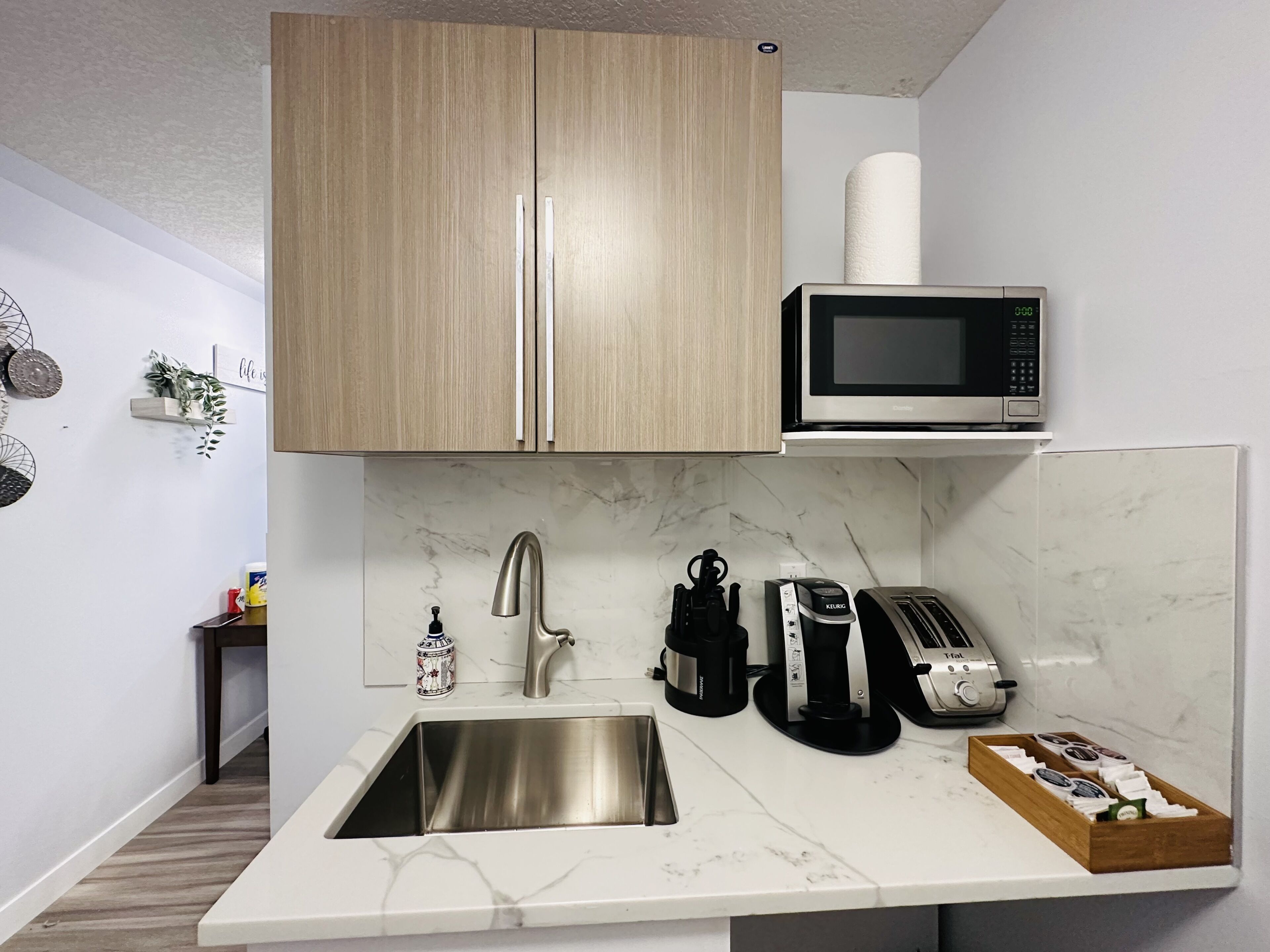 A compact kitchen with wooden cabinets, a stainless steel sink, a microwave, a toaster, and a coffee maker.