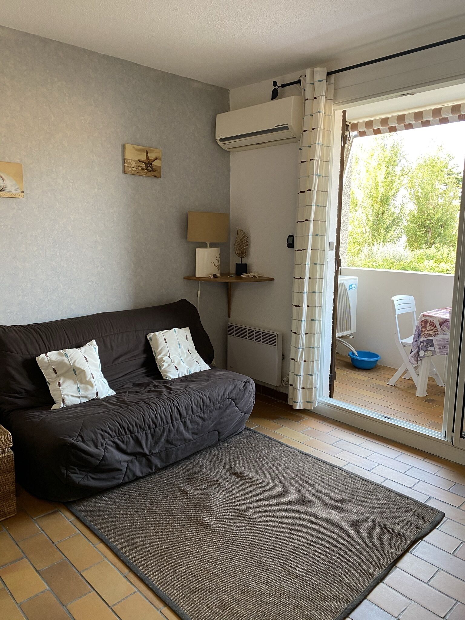 En hyggelig stue med en mørk sofa, et lille bord med en lampe og udsigt gennem en skydedør til et udendørs område med et bord og stole.
