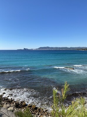A coastal view with clear blue water, rocky shoreline, and distant hills.
