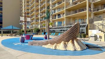 Area piscina con scivolo a forma di relitto, lettini e un edificio a più piani sullo sfondo.