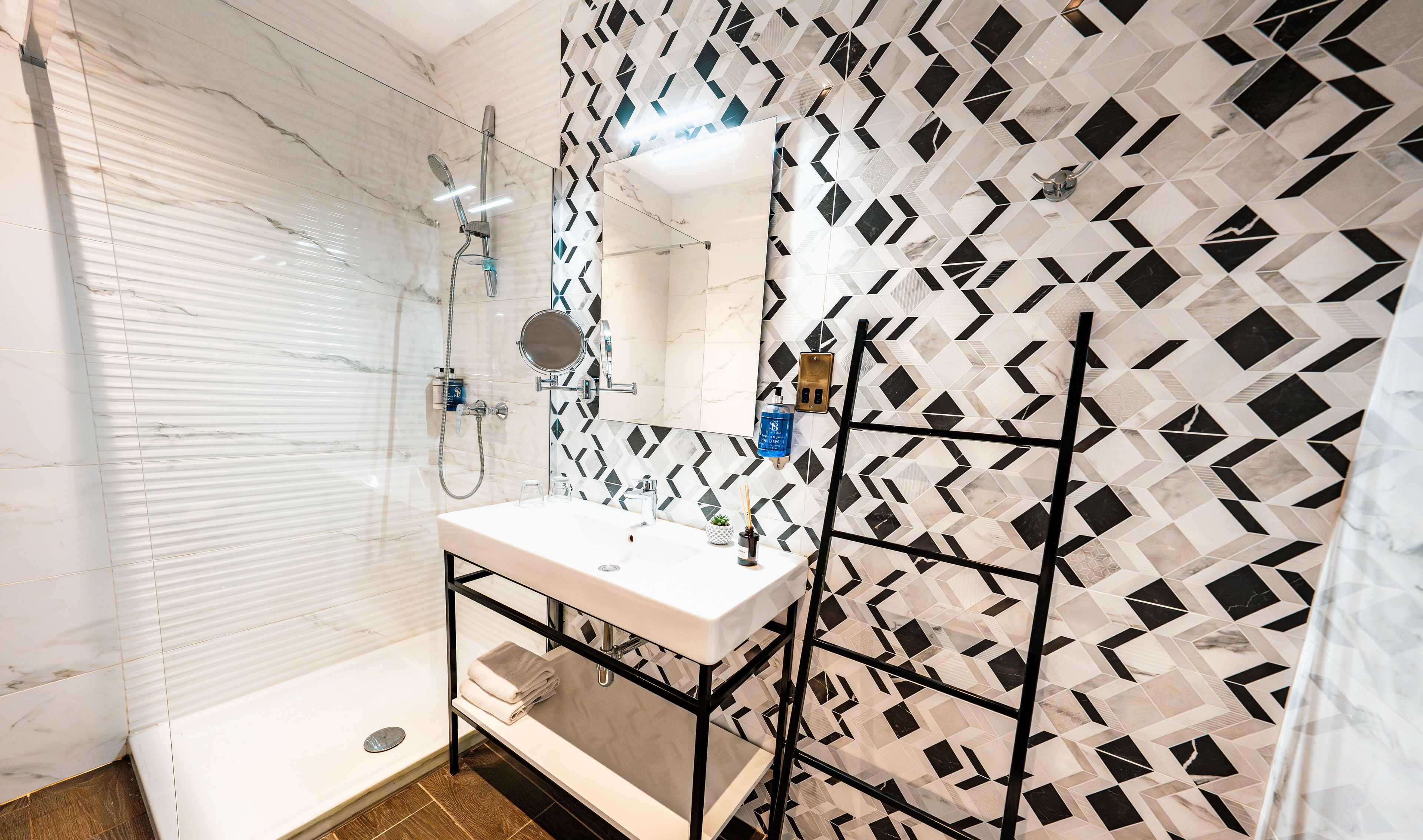 Un baño moderno con ducha de vidrio, un lavamanos blanco con estructura metálica negra y pared con diseño.
