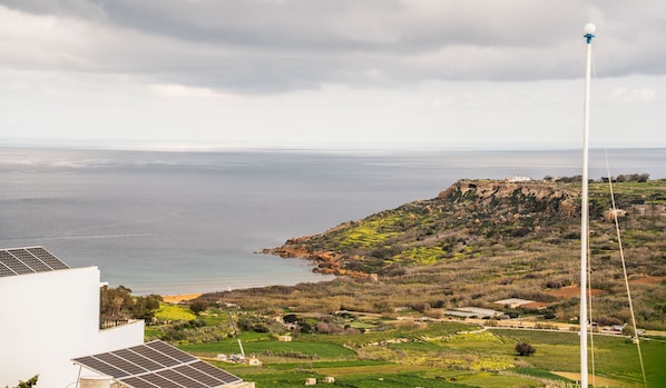 Paisaje costero con una turbina eólica, paneles solares y vistas al mar y colinas.