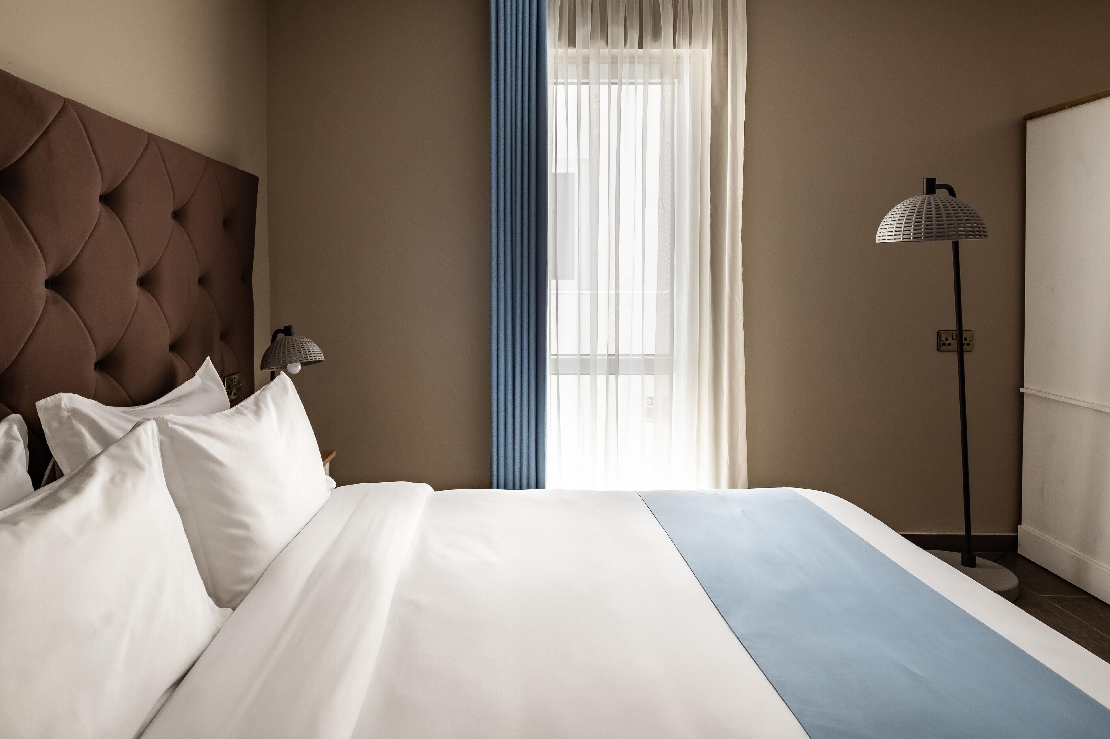 Une chambre d’hôtel avec un grand lit, une tête de lit, une lampe de chevet, une fenêtre avec des rideaux et un lampadaire.
