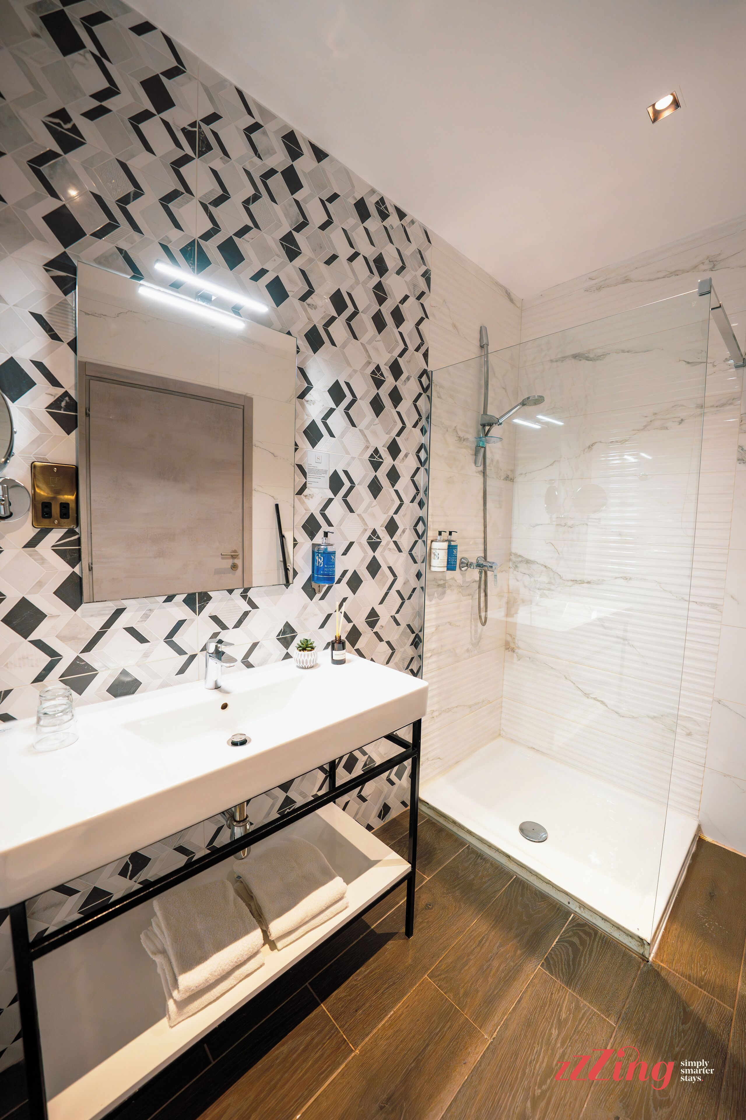 A modern bathroom with a geometric patterned wall, a white sink with a mirror, a glass shower enclosure, and a wooden floor.