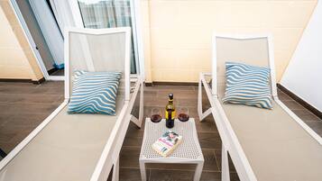 Een balkon met twee witte lounge fauteuils, elk met een blauw-wit gestreept kussen. Op een klein tafeltje tussen de stoelen staan een fles wijn en twee glazen.