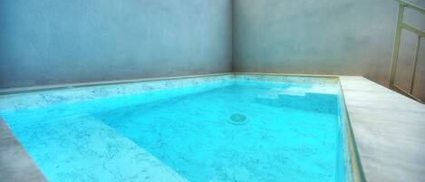 A rectangular swimming pool with a marble-like floor and walls, surrounded by a simple, light-colored structure.