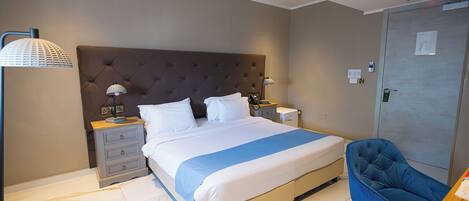 A modern hotel room with a large bed, a chest of drawers, a desk, a blue armchair, and a wardrobe.