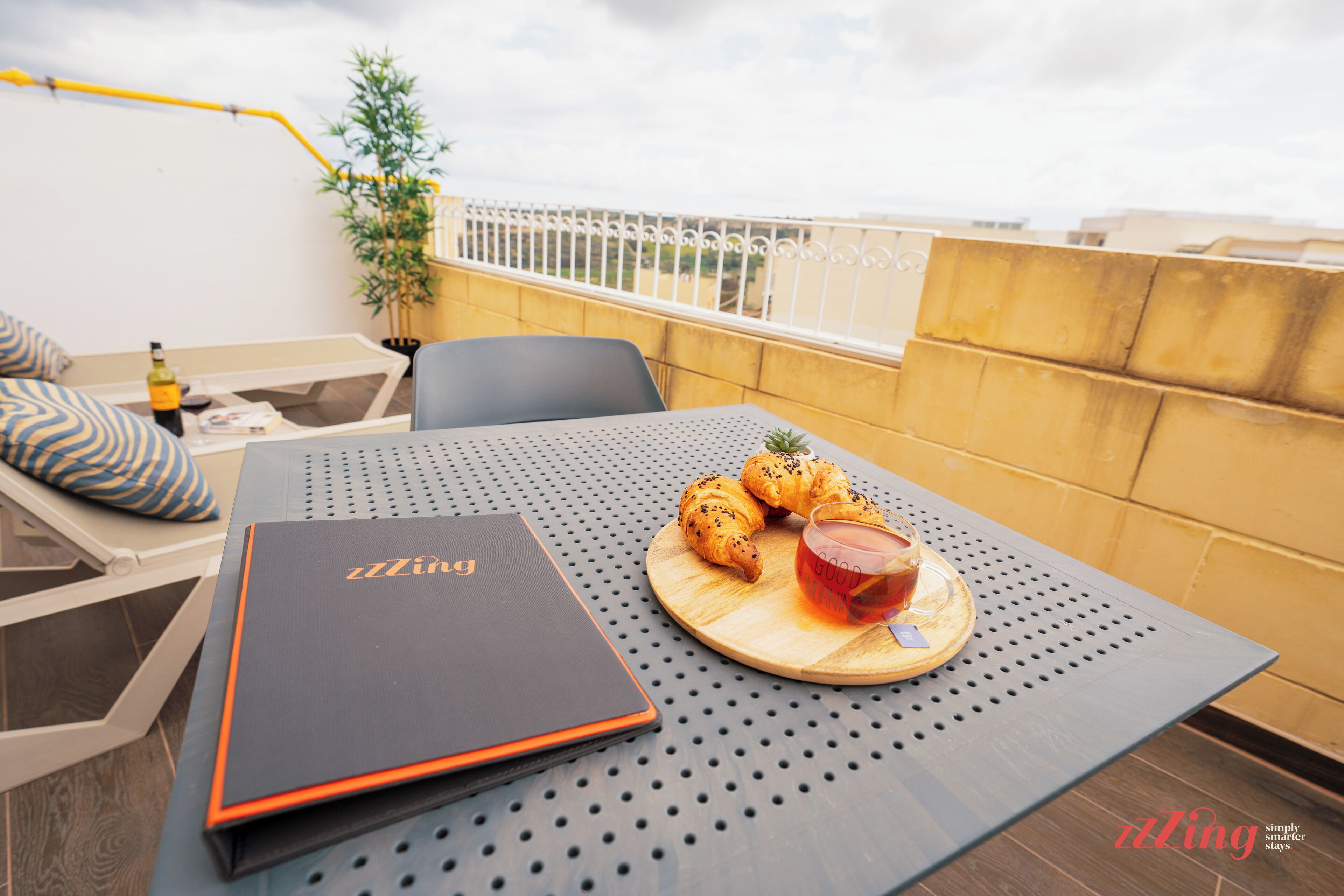 A table with a laptop, croissants, and a glass of red beverage on a balcony with a view of buildings.