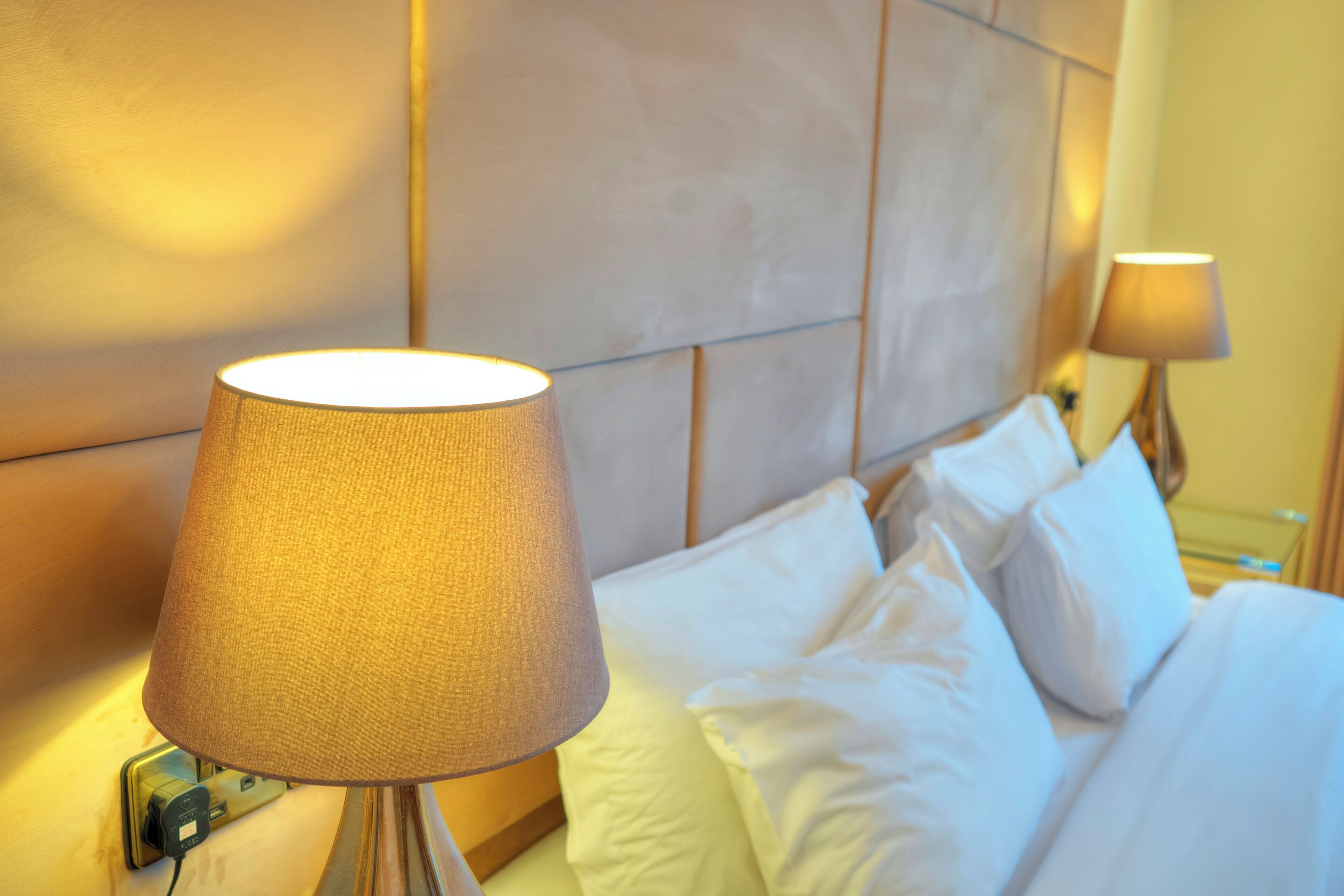 Hotellihuone, jossa on sänky, valkoiset vuodevaatteet ja seinään kiinnitetty lamppu.