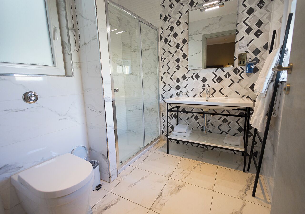 A bathroom with a toilet, a glass shower enclosure, a sink with a black metal shelf, and a patterned wall.