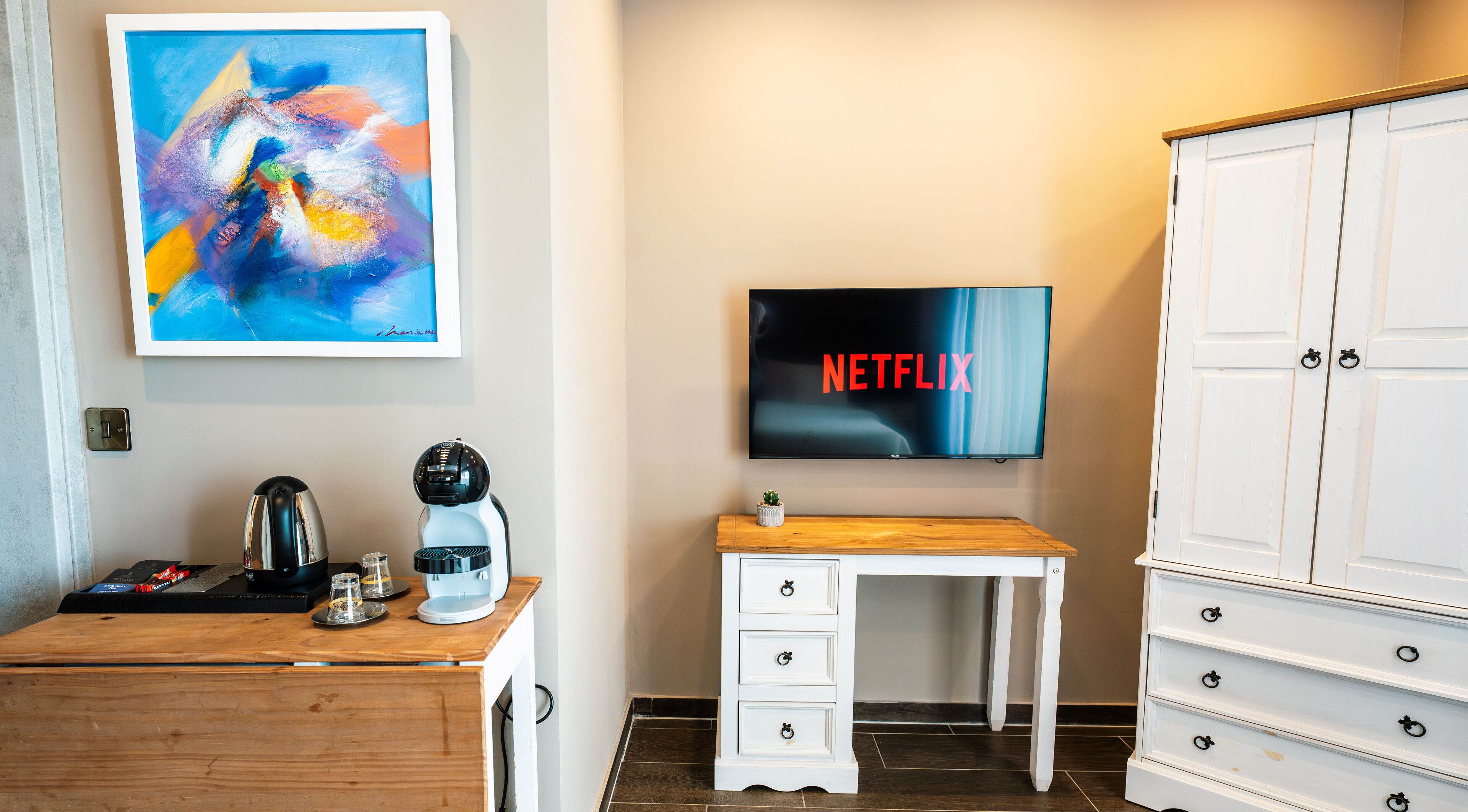 A hotel room with a wooden desk, a coffee maker, a TV displaying the Netflix logo, a white dresser, and a painting on the wall.