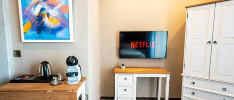 A hotel room with a wooden desk, a coffee maker, a TV displaying the Netflix logo, a white dresser, and a painting on the wall.