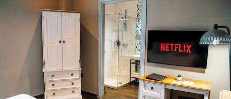 Un baño moderno con cabina de ducha de vidrio, un armario blanco, tocador con lavabo y un televisor de pantalla plana mostrando el logotipo de Netflix.