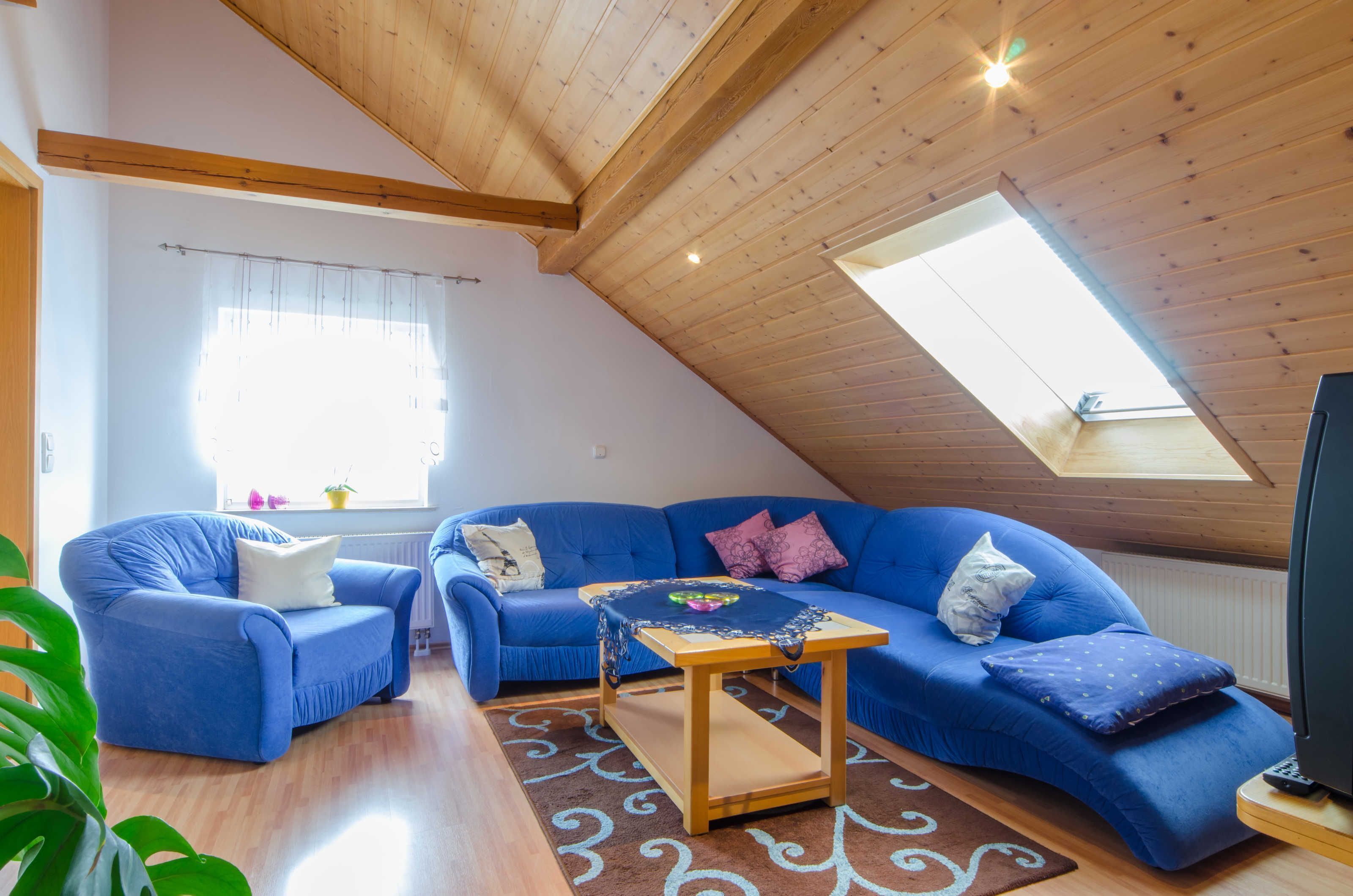 Una sala de estar acogedora con techo inclinado, un sofá azul, una mesa de centro de madera y un amplio ventanal que proporciona luz natural.