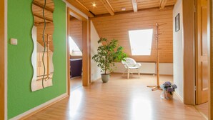 Une pièce avec un plancher en bois, un mur vert orné d’un miroir, une plante en pot, une chaise blanche et un plafond en bois.