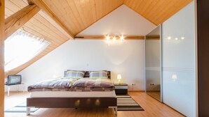 Une chambre avec un plafond en pente, un lit avec une literie à motifs, une table de chevet avec une lampe, un téléviseur posé sur un petit meuble et une porte-fenêtre coulissante.