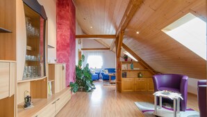 Un loft spacieux avec des poutres en bois, une lucarne, un canapé bleu, un fauteuil violet et un meuble en bois contenant de la verrerie.