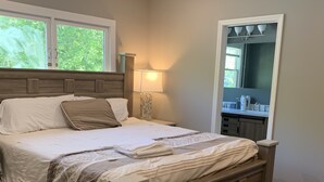A bedroom with a wooden bed, a nightstand with a lamp, a window with a view of trees, and a bathroom with a mirror and sink. - Rare Find! 6 Bedroom Duplex, Close to Downtown! (Atlanta)