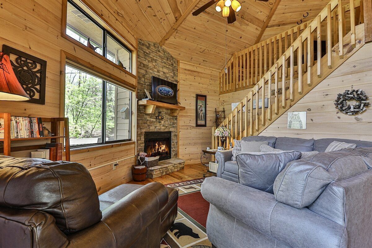 A cozy cabin interior with a fireplace, wooden walls, a staircase, and a large gray sofa.
