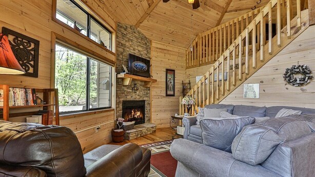 A cozy cabin interior with a fireplace, wooden walls, a staircase, and a large gray sofa. - A Tucked Away Lodge | Ellijay, GA (Ellijay)