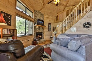A cozy cabin interior with a fireplace, wooden walls, a staircase, and a large gray sofa.