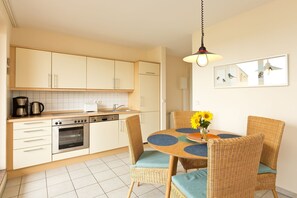 Une cuisine moderne avec des meubles blancs, un four encastré et un coin repas agrémenté de chaises en osier et d’une table ronde.