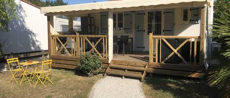 Un'area esterna riparata con un pavimento in legno, un tavolino con sedie e un vialetto sterrato che conduce a un edificio.