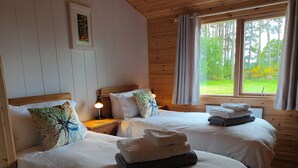 Una habitación acogedora con dos camas, paredes de madera, una ventana con vistas a la vegetación y una mesita de noche con una lámpara.