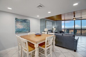 A dining area with a wooden table and white chairs, a sofa, and a view of the ocean through large windows.