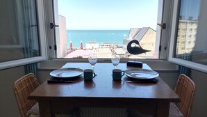 A dining table with plates, a wine glass, and a cup, set against a window overlooking a beach with umbrellas and a swan statue. - THE HORIZON (Fécamp)