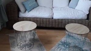 A white wicker sofa with multiple cushions, two round tables with wireframe bases, and a patterned rug. - THE HORIZON (Fécamp)