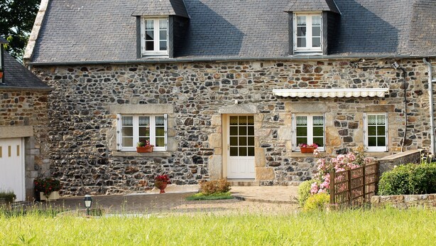 A stone house with a sloped roof, two windows with white frames, a front door, and a small porch area with potted plants. - Longère Bretonne Dans un Écrin de Verdure (Pléboulle)