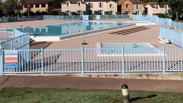 A swimming pool with a blue fence, surrounded by a grassy area and a row of houses in the background. - House in vacation village (Monflanquin)
