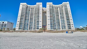 Een hoog hotel met meerdere balkons, een centrale ingang en een zandstrand op de voorgrond.
