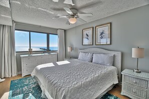 A bedroom with a large bed, a ceiling fan, two framed pictures, a nightstand, and a view of the ocean. - Ocean Bay Club 810 | Oceanfront Four Bedroom Condo (North Myrtle Beach)