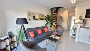 Una sala moderna con un sofá gris, una mesa de centro, una silla, una lámpara, un televisor y una zona de cocina con un microondas.