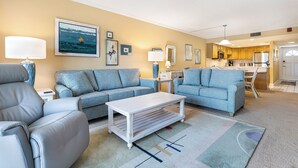 Ein Wohnzimmer mit Sofa, Sessel und Couchtisch, im Hintergrund eine Küche und ein Gemälde an der Wand.